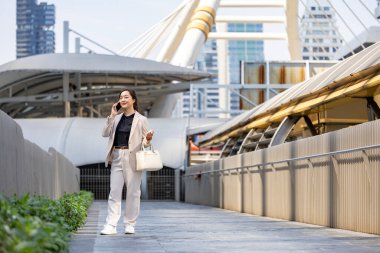 Şehir merkezindeki gökdelen caddesi modern ofisinde iş iletişimi ve kentsel yaşam tarzı üzerine yürürken cep telefonu kullanan lüks genç Asyalı CEO kadın girişimci.