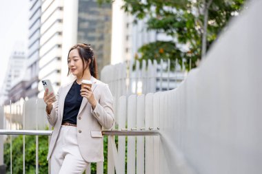 Şehir merkezindeki gökdelen caddesi modern ofisinde iş iletişimi ve kentsel yaşam tarzı kavramı üzerine yürürken cep telefonu kullanan lüks genç Asyalı CEO kadın girişimci.