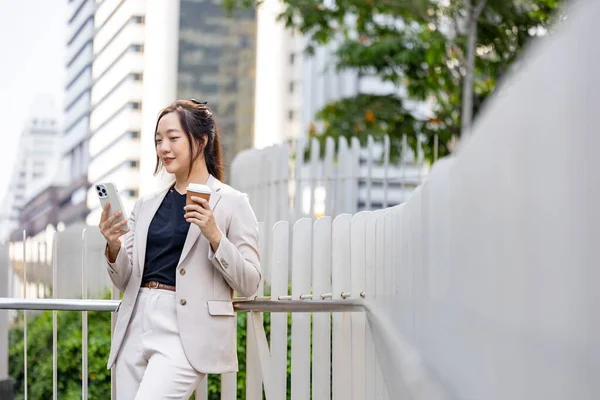 Şehir merkezindeki gökdelen caddesi modern ofisinde iş iletişimi ve kentsel yaşam tarzı kavramı üzerine yürürken cep telefonu kullanan lüks genç Asyalı CEO kadın girişimci.