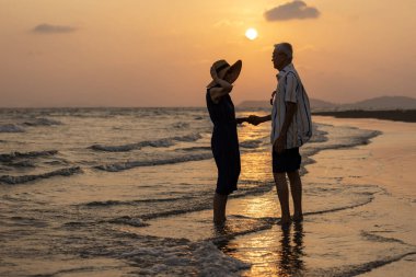 Asyalı yaşlı çiftin silueti yaz mevsiminde günbatımı deniz sahilinde tatillerinin tadını çıkarıyor sağlıklı bir ilişki ve emeklilik yıllarında mutlu bir evlilik hayatı için.