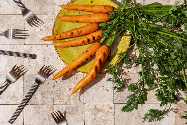 Bunch of grilled carrot roots with green leaves and salt flakes on a green craft plate on stone tales of earth tones background with six forks top view. Healthy eating and dieting food concept.