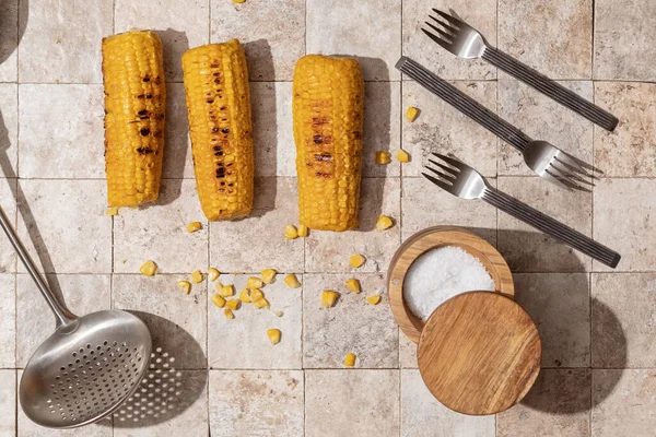 Delicious organic homemade grilled corn cobs with three forks, salt cellar, slotted spoon on earth toned stone tales background top view