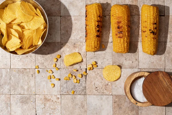 Delicious organic homemade grilled corn cobs and bowl of nacho chips with salt cellar on earth toned stone tales background top view