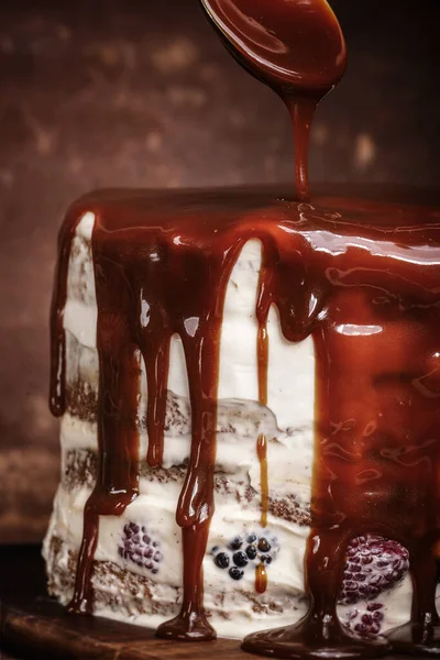 Pouring caramel sauce with spoon onto caramel cake with wild berries, cream on brown background