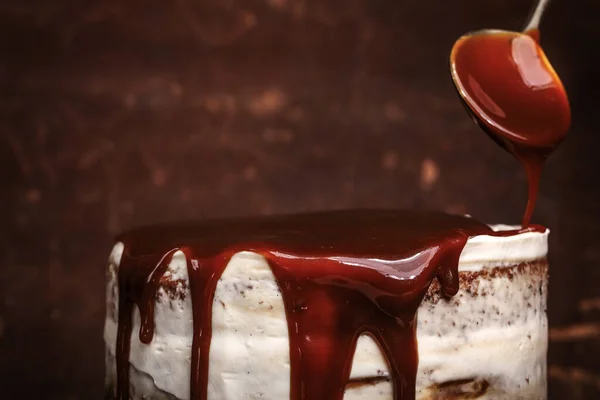 Pouring caramel sauce with spoon onto caramel cake with wild berries, cream on brown background