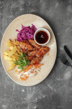 Izgara tavuk kanadı ve sebzeli fırında patates. Gri kil arka planda, sos ve çatal bıçaklı.