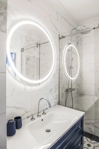 Modern banyo içi koyu mavi ahşap bir raf, beyaz lavabo ve LED ışıklı büyük yuvarlak ayna.. 