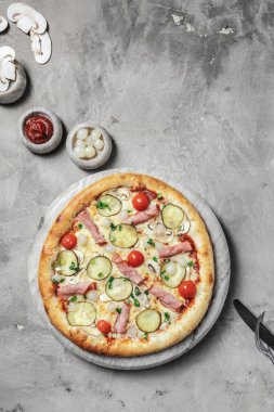 Jambonlu, kiraz domatesli, mozarellalı, mantarlı, yeşil soğanlı ve mermer tepside salatalık turşulu lezzetli pizza. Gri taşlı arka planda pizza malzemeleriyle süslenmiş. 