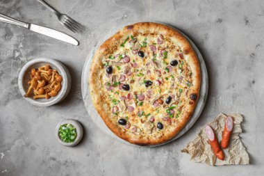 Sosisli lezzetli pizza, mozzarella, marine edilmiş mantar, siyah zeytin, mermer tepside yeşil soğan. Gri taşlı arka planda pizza malzemeleriyle süslenmiş. 