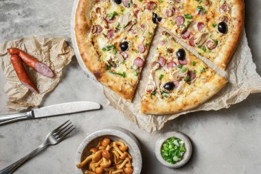 Sosisli lezzetli pizza, mozzarella, marine edilmiş mantar, siyah zeytin, mermer tepside yeşil soğan. Gri taşlı arka planda pizza malzemeleriyle süslenmiş. 