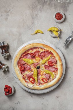 Jambonlu lezzetli pizza, mozzarella, mermer tepside marine edilmiş biber. Gri taşlı arka planda pizza malzemeleriyle süslenmiş. 