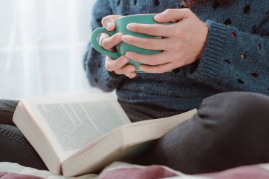 Bir kadının bir fincan sıcak kahve içerken, rahat bir şekilde kitap okurken yakın çekimi.