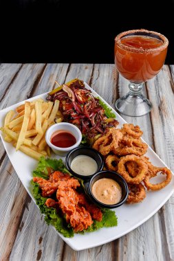 Restoran masasında bir ön plan vardır. Kızarmış patates, kızarmış soğan, tavuk kanadı ve üç farklı Meksika sosu içeren büyük bir tabak abur cubur.