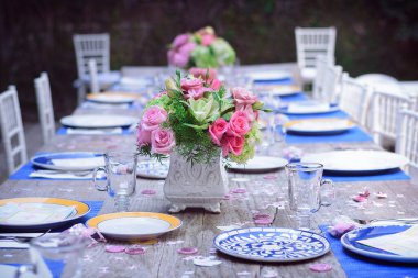 Bahçedeki masada mavi ve sarı porselen tabaklar, boş kristal bardaklar ve ortasında pembe güller ve beyaz sandalyeler ile bir çiçek düzenleme, arka planda bulanıklık var.