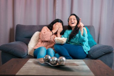 Evde film izleyen bir anne ve kızın yatay görüntüsü, patlamış mısır yiyorlar, kızı korkuyor ve annesi yaptığı yüz ifadelerine gülüyor.