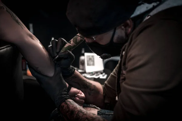 A horizontal shot of a tattoo artist doing a tattoo to a woman on her arm, the tattoo artist is very concentrated operating the tattoo machine