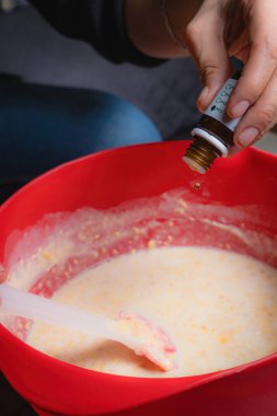 Evde, bir kadının elleri kırmızı bir kapta turuncu esanslı organik sabun karıştırırken görülebilir. Yüzde yüz doğal.