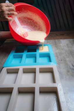 Evde, doğal sabunun silikon kaplara döküldüğü kırmızı bir kap tutan bir kadının ellerini görebilirsiniz. Sabun% 100 organik ve ev yapımı..