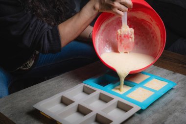 Evde, doğal sabunun silikon kaplara döküldüğü kırmızı bir kap tutan bir kadının ellerini görebilirsiniz. Sabun% 100 organik ve ev yapımı..