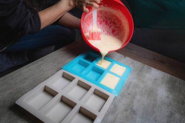 Evde, doğal sabunun silikon kaplara döküldüğü kırmızı bir kap tutan bir kadının ellerini görebilirsiniz. Sabun% 100 organik ve ev yapımı..