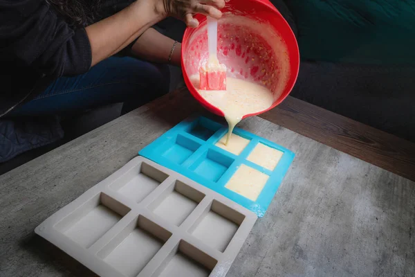 Evde, doğal sabunun silikon kaplara döküldüğü kırmızı bir kap tutan bir kadının ellerini görebilirsiniz. Sabun% 100 organik ve ev yapımı..