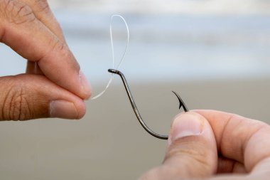 Beyaz bir adamın elleri denizin önündeki siyah bir oltanın gözüne naylon bir olta atıyor. Kıyı balıkçılığına, spora ve güneşli bir hafta sonunda ailenle geçirdiğin boş zamanlardan bahset..