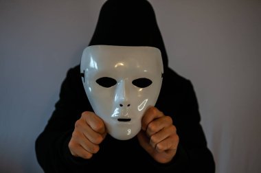 Siyah kapüşonlu bir adam siyah arka planda isimsiz beyaz bir maske tutuyor.