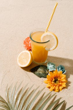 beach sand background with a fruit juice and lemon pieces, some colorful flowers and a coconut tree leaf. summer and holiday concept. vertical image with copy space.