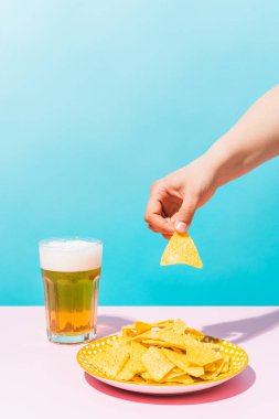 Bilinmeyen bir kadının eli, mavi ve pembe arka plandaki biranın yanındaki tabaktan nacho alıyor. Yemek, Meksika ve yaz konsepti.