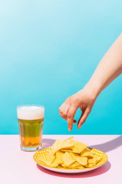 Mavi ve pembe arka planda biranın yanındaki tabaktan nacho almak üzere olan bilinmeyen bir kadının eli. Yemek, Meksika ve yaz konsepti.
