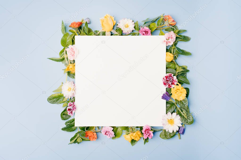 Blank paper card with square floral frame of assorted flowers on blue pastel background. Top view mockup with copy space.