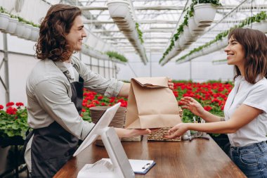 Erkek çiçekçinin portresi, önlük takıyor ve bahçe merkezindeki veya çiçekçideki kadına hediye veriyor.