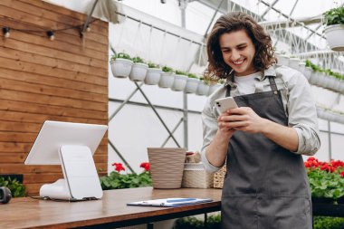 Çiçekçide çalışırken gülümseyen ve akıllı telefon kullanan genç bir çiçekçi.