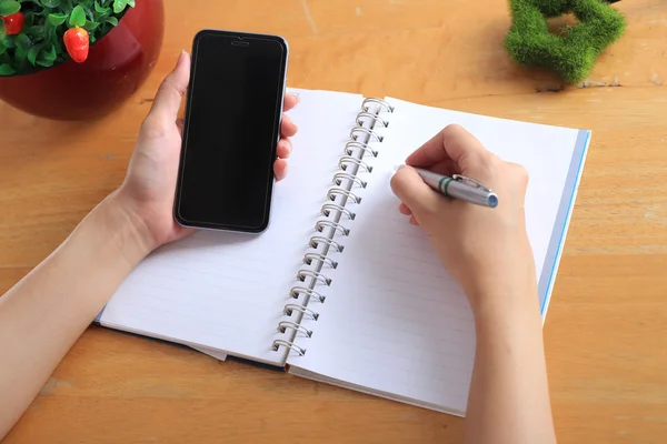 Escritório da mulher escrever notas e usar telefone inteligente na mesa de madeira . — Fotografia de Stock
