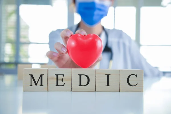 Close-up doctor in uniform wearing a face mask with a stethoscope and hold red heart with wood cubes word MEDIC.