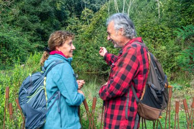 Mutlu yaşlı çift, yemyeşil bir patika boyunca birlikte yürüyüş yapıyor. Huzurlu bir gölet kenarında, gülümsüyor, sırt çantaları taşıyor ve aktif, sağlıklı emekliliğin ve doğal huzurun tadını çıkarıyor.