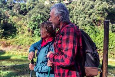 Üst düzey bir çift, aktif ve sağlıklı bir emeklilik yolculuğu sırasında doğayla bağlantılı güneşli yeşil bir manzaranın ve suyun tadını çıkarmak için duraklıyor.