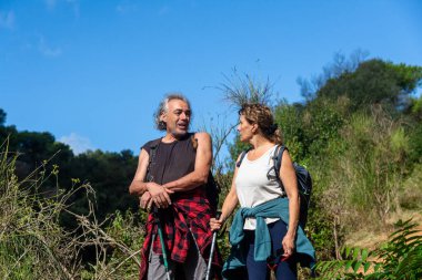 Üst düzey bir çift, iletişim kurarken, aktif yaşam tarzı ve birlikteliği temsil ederken, yürüyüş direkleri ile doğal bir arazide yürüyüş yapıyor.