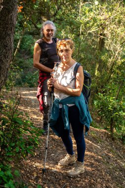 Aktif kıdemli çift orman patikalarını keşfediyor, yürüyüşe ara verip kameraya gülümsüyor, güneşli bir günde birlikte açık hava macerasının tadını çıkarıyorlar.