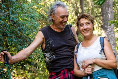 Kıdemli çift gülümsüyor ve gülüyor, yürüyüş direkleri tutuyorlar, canlı yeşil orman ortamında aktif olarak sağlıklı bir yaşam tarzına giriyorlar, birlikte bir anlık mutluluğu paylaşıyorlar.