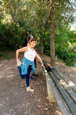 Yaşlı bir kadın, yemyeşil ağaçların arasında eşiyle yürüyüş yaparken bankta bacağını esnetiyor. Aktif yaşlanma ve açık hava yaşam tarzını destekliyor.
