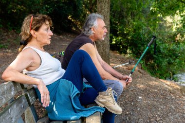 Yaşlı bir adam ve kadın, sakin bir orman ortamında ahşap bir bankta mola verir, doğa ile çevrili aktif bir doğa yürüyüşünden sonra yürüyüş direkleri ile dinlenir.