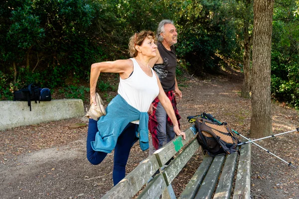 Kıdemli bir kadın, kocası yakınlarda dururken parktaki bankta bacaklarını esnetiyor. İkisi de bir orman ortamında açık hava aktivitesine hazırlanıyor. Aktif yaşam tarzını destekliyor.