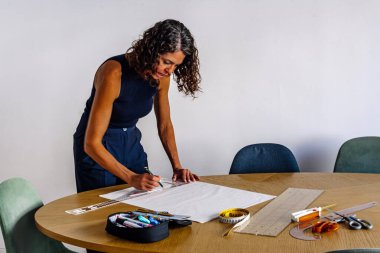 Kadın moda tasarımcısı, ahşap bir masada çizim araçları üzerinde çalışıyor. Bağımsız işi için yeni tasarımlar geliştirme sürecine odaklanıyor.