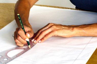 Beyaz kağıda kalem ve cetvelle moda tasarımı çizen kadın elleri, stüdyo çalışma alanındaki hatlara ve ölçümlere odaklanmış, el işi ve beceri sergiliyor.