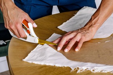 Kadınların elleri, beyaz tekstil kumaşı ahşap bir masa üzerinde kesiyor. Kahverengi kağıt desenini takip ediyor. Moda oluşumunu ve bağımsız tasarım çalışmalarını temsil ediyor.