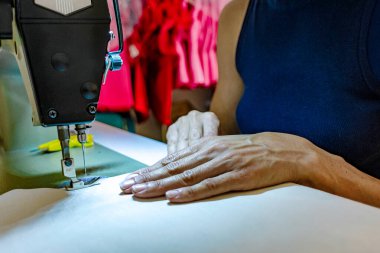 Stüdyoda dikiş makinesi işleten yetenekli bir kadın tasarımcı. Odaklanmış elleri ve detaylara dikkat eden el yapımı bir giysi yapıyor. Bağımsız terziliği ve küçük işletmeleri temsil ediyor.