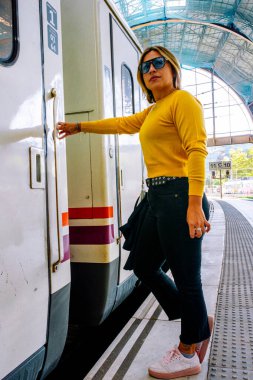 Güneş gözlüklü ve sarı tişörtlü genç bir kadın, güneşli bir istasyonda trene biniyor, özgüven ve seyahat tutkusu ile tek başına yaz yolculuğuna çıkıyor.