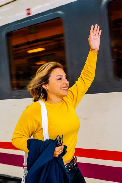 Bir istasyon platformunda duran, gülümseyen ve elini sallayan genç bir kadın, seyahat duygularını ifade ediyor, elveda ve hareket eden beyaz ve kırmızı bir tren arkasından hızla geçerken