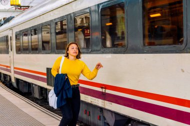 Sıradan bir ceket ve çanta giymiş genç bir kadın kalabalık bir şehir tren platformunda duruyor, tek başına yolculuk ve maceraya hazırlanan bir banliyö trenine binmeyi bekliyor.